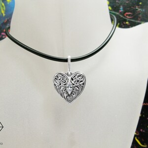 May include: A silver heart pendant necklace with a clear crystal in the center. The pendant has a filigree design and is hanging from a black cord.