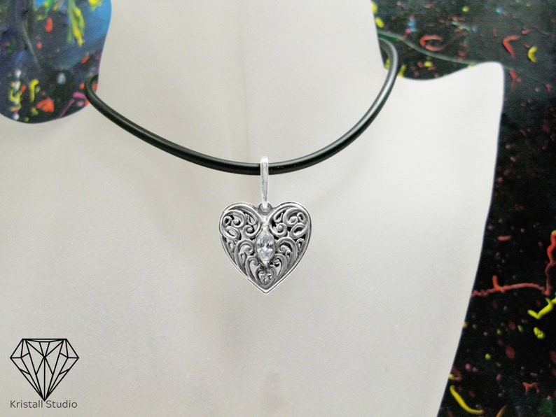 May include: A silver heart pendant necklace with a clear crystal in the center. The pendant has a filigree design and is hanging from a black cord.