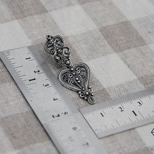 May include: A silver filigree heart-shaped earring with intricate details. The earring is measured against a ruler, showing its size.
