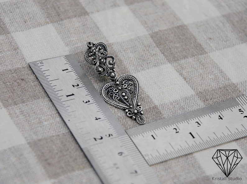 May include: A silver filigree heart-shaped earring with intricate details. The earring is measured against a ruler, showing its size.
