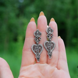 May include: A pair of silver filigree earrings with a heart-shaped design. The earrings are dangling and have a delicate, intricate pattern.