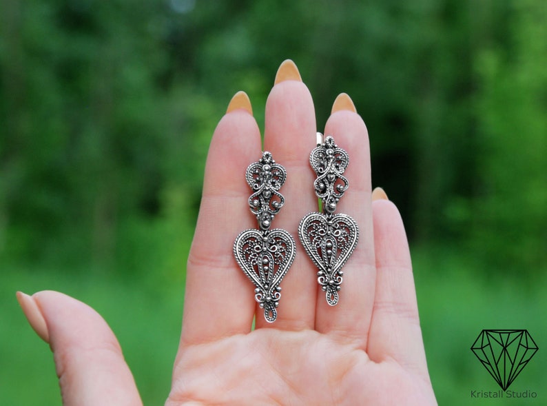 May include: A pair of silver filigree earrings with a heart-shaped design. The earrings are dangling and have a delicate, intricate pattern.