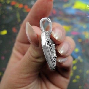 May include: Silver heart-shaped pendant with intricate details and a loop at the top for attaching a chain. The pendant is held in a hand.