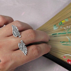 May include: Two silver rings shaped like angel wings. The rings are worn on the index and middle fingers of a hand.