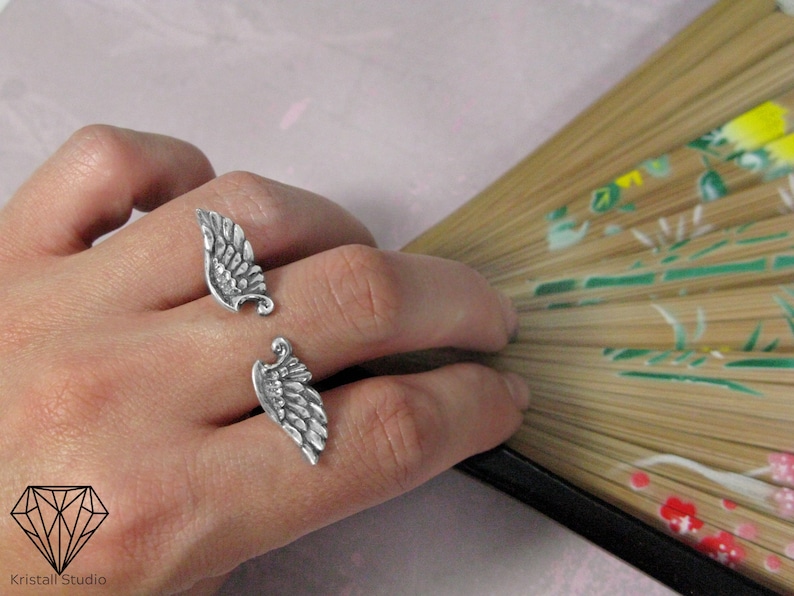 May include: Two silver rings shaped like angel wings. The rings are worn on the index and middle fingers of a hand.