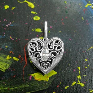 May include: A silver filigree heart pendant with a clear crystal in the center. The pendant is on a black background with colorful paint splatters.