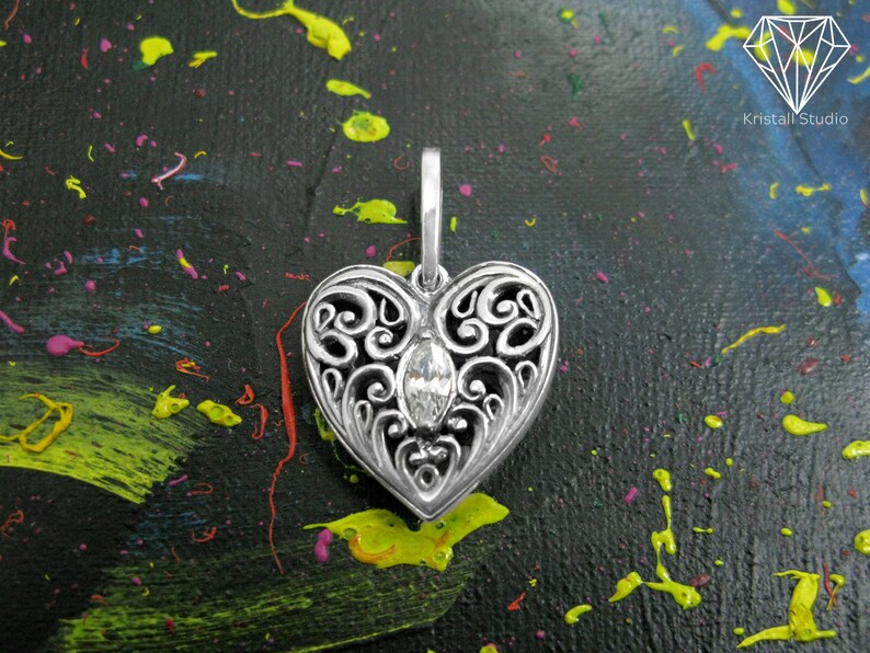May include: A silver filigree heart pendant with a clear crystal in the center. The pendant is on a black background with colorful paint splatters.