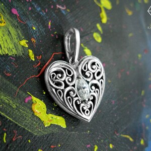 May include: A silver heart-shaped pendant with an intricate filigree design and a clear gemstone in the center. The pendant is on a black background with colorful paint splatters.