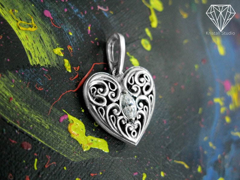 May include: A silver heart-shaped pendant with an intricate filigree design and a clear gemstone in the center. The pendant is on a black background with colorful paint splatters.