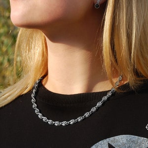 May include: A silver chain necklace with a unique design featuring small, interconnected circles. The necklace is worn by a person with long blonde hair and a black shirt.