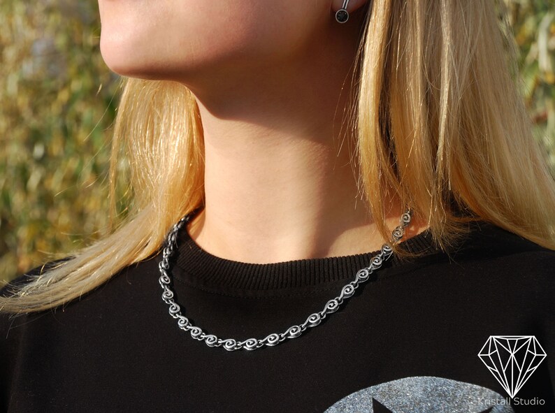 May include: A silver chain necklace with a unique design featuring small, interconnected circles. The necklace is worn by a person with long blonde hair and a black shirt.