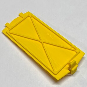 May include: A yellow plastic cover with a rectangular shape and a series of internal ridges. The cover has a small tab on the right side and the text "1991" is visible on the surface.