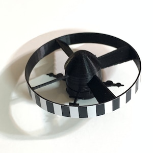 May include: A black and white striped ring with a black propeller in the center. The propeller has four blades and is attached to a black hub.