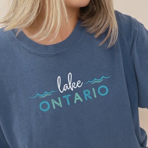 May include: A blue t-shirt with the text "lake ONTARIO" in white and blue lettering with wavy lines above and below the text. The shirt is worn by a person sitting down with white pants that say "DENIM" on the leg.