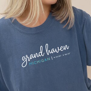 May include: A blue denim t-shirt with the text "grand haven MICHIGAN | N 43.06° | W 86.23°" printed on the front.