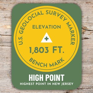 Könnte beinhalten: Ein runder Aufkleber in Grün und Gelb mit dem Text "U.S. Geological Survey Marker" und "Elevation 1,803 FT. Bench Mark". Der Aufkleber hat auch den Text "High Point" und "Highest Point in New Jersey".