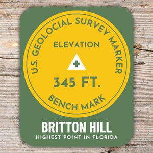 Könnte beinhalten: Ein runder Aufkleber in Grün und Gelb mit dem Text "U.S. Geological Survey Marker" und "Elevation 345 FT. Bench Mark". Der Aufkleber trägt außerdem den Text "Britton Hill Highest Point in Florida".
