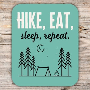 May include: A teal blue coaster with the text "HIKE, EAT, sleep, repeat." in black. The coaster features a black silhouette of a campfire, tent, and pine trees against a light blue sky with a crescent moon.