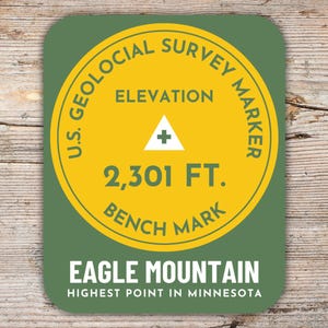 Könnte beinhalten: Ein runder Aufkleber in Grün und Gelb mit dem Text "U.S. Geological Survey Marker" und "Elevation 2,301 FT. Bench Mark". Unter dem Aufkleber steht der Text "Eagle Mountain Highest Point in Minnesota".