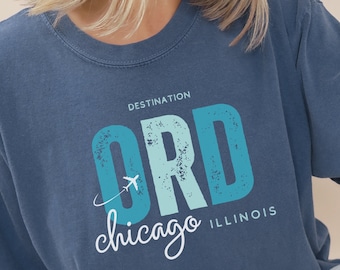 May include: A blue t-shirt with a distressed graphic design that says "DESTINATION ORD chicago ILLINOIS" in white and blue lettering. The shirt is made of denim.