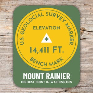 Könnte beinhalten: Ein runder Aufkleber in Grün und Gelb mit dem Text "U.S. Geological Survey Marker" und "Elevation 14,411 FT. Bench Mark". Der Aufkleber trägt außerdem den Text "Mount Rainier Highest Point in Washington".