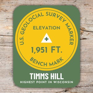 Può includere: Un adesivo rotondo verde e giallo con il testo "U.S. Geological Survey Marker" e "Elevation 1,951 FT. Bench Mark". L'adesivo ha anche il testo "Timms Hill Highest Point in Wisconsin".
