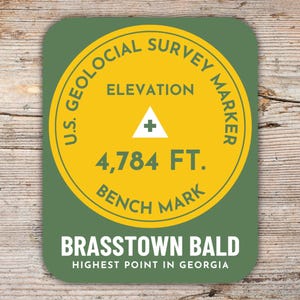 Könnte beinhalten: Ein runder Aufkleber in Grün und Gelb mit dem Text "U.S. Geological Survey Marker" und "Elevation 4,784 FT. Bench Mark". Der Aufkleber trägt außerdem den Text "Brasstown Bald Highest Point in Georgia".