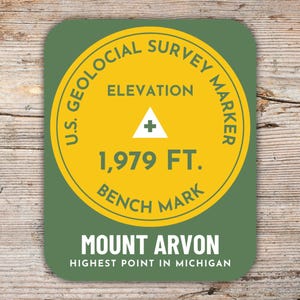 Könnte beinhalten: Ein runder Aufkleber in Grün und Gelb mit dem Text "U.S. Geological Survey Marker" und "Elevation 1,979 FT. Bench Mark". Der Aufkleber trägt außerdem den Text "Mount Arvon Highest Point in Michigan".