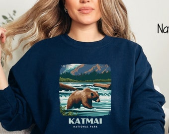 May include: A navy blue sweatshirt with a graphic of a brown bear in Katmai National Park. The graphic is a vintage-style illustration of a bear standing in a river with mountains in the background.