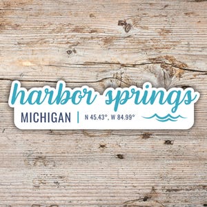 May include: A white sticker featuring the words "harbor springs" in teal script. Beneath, it states "MICHIGAN | N 45.43°, W 84.99°" in a smaller font. Two teal wave graphics are at the bottom. The background is a light brown wooden surface.