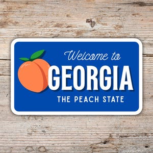 May include: A rectangular sticker with a blue background and white border. The sticker features a peach graphic and the text "Welcome to GEORGIA THE PEACH STATE" in white. The sticker is placed on a wooden surface.