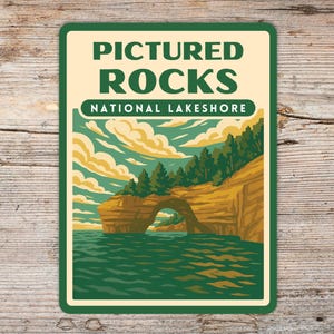 May include: Rectangular sign featuring the text "PICTURED ROCKS NATIONAL LAKESHORE" above an illustration of a rock formation with an arch over water. The sign has a green border and a cream-colored background.