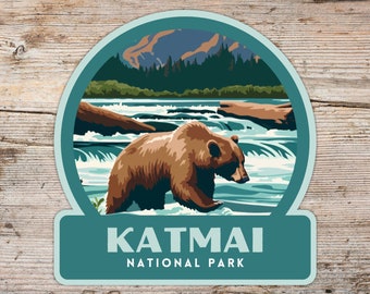May include: A blue and green illustrated sticker featuring a brown bear standing in a river with a mountain range in the background. The text "Katmai National Park" is printed below the bear.