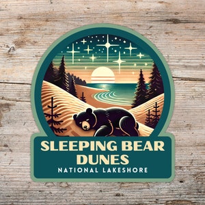 May include: A green and blue circular sticker featuring a black bear sleeping on a sand dune. The sticker displays the text "Sleeping Bear Dunes National Lakeshore" in white.