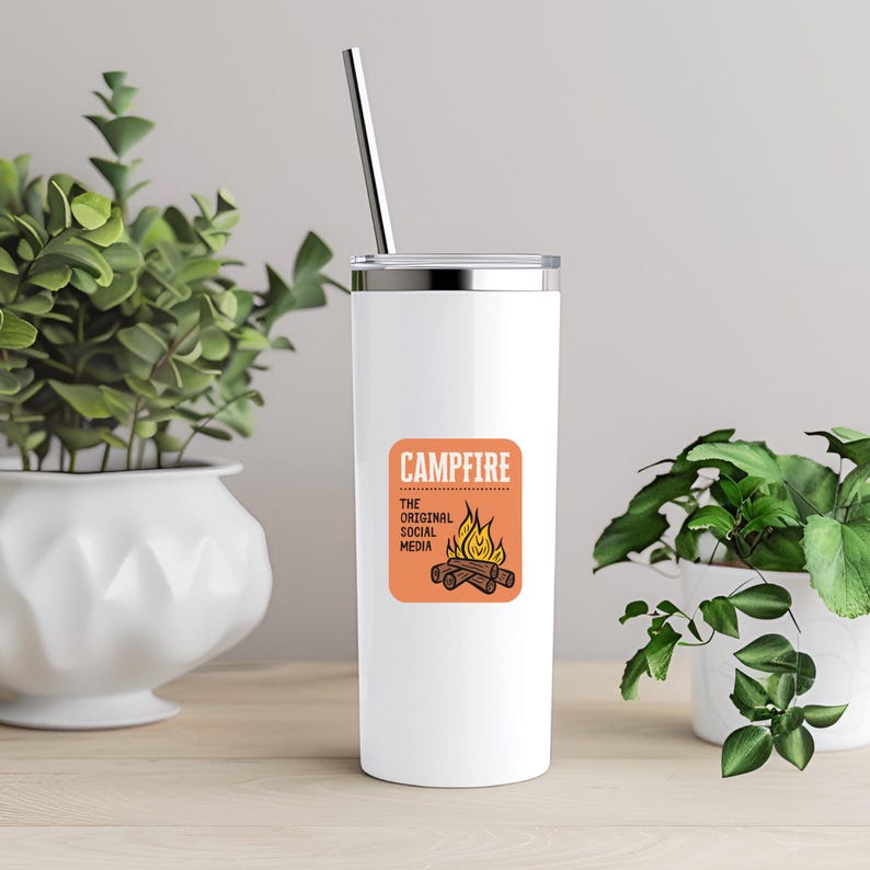 May include: A white insulated tumbler with a silver lid and straw. The tumbler features a square orange graphic with the word "CAMPFIRE" and a campfire illustration. The text "THE ORIGINAL SOCIAL MEDIA" is below the word "CAMPFIRE".