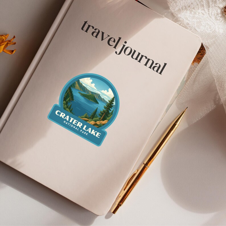 May include: A cream-colored travel journal with the words "travel journal" printed in black. A Crater Lake National Park sticker is affixed to the journal. A gold pen rests on the journal. The sticker features a blue lake scene with trees and mountains.