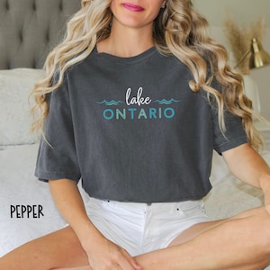May include: A dark gray t-shirt with the text "lake ONTARIO" in blue and white lettering. The text is surrounded by a wavy line design.