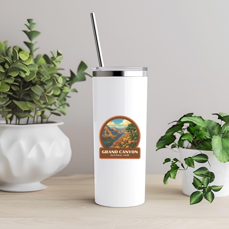 May include: A white travel tumbler with a silver lid and a metal straw. The tumbler features a circular sticker with a Grand Canyon National Park design. The design shows a canyon landscape with a blue sky and the words "Grand Canyon National Park".