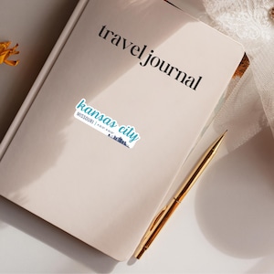 Puede incluir: Un diario de viaje beige con el texto "travel journal" impreso en la portada. Una pegatina con el texto "kansas city" y las coordenadas "39.0997&deg; N, 94.5786&deg; W" est&aacute; en la portada.