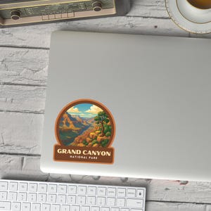 May include: A circular sticker featuring a scenic illustration of the Grand Canyon National Park. The design includes a canyon landscape with a river, a tree, and the text "GRAND CANYON NATIONAL PARK" in a brown rectangular banner. The sticker is on a gray surface.
