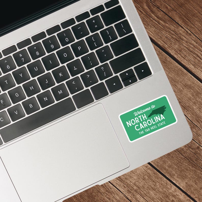 Puede incluir: Una pegatina verde con el texto "Welcome to NORTH CAROLINA THE TAR HEEL STATE" y una silueta de Carolina del Norte est&aacute; adherida a un port&aacute;til. El port&aacute;til est&aacute; sobre una superficie de madera.