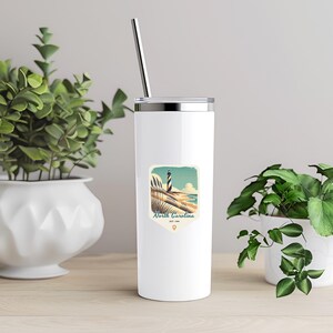May include: White stainless steel tumbler with a straw. The tumbler has a graphic of a lighthouse on a beach with the text "North Carolina Est. 1999".