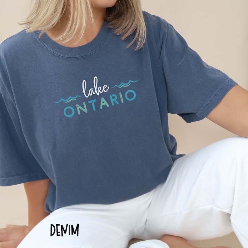 May include: A blue t-shirt with the text "lake ONTARIO" in white and blue lettering with wavy lines above and below the text. The shirt is worn by a person sitting down with white pants that say "DENIM" on the leg.