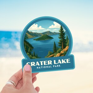 May include: A sticker featuring a scenic illustration of Crater Lake National Park. The design includes a blue lake, green island, and surrounding mountains, with the text "CRATER LAKE NATIONAL PARK" in white.