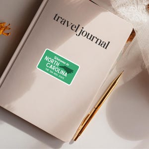 Puede incluir: Un diario de viaje color crema con las palabras "travel journal" impresas en negro. Una pegatina verde y blanca que dice "Welcome to North Carolina, The Tar Heel State" est&aacute; adherida al diario. Un bol&iacute;grafo dorado descansa sobre el diario.