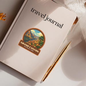 May include: A cream-colored travel journal with the words "travel journal" printed in black. A Grand Canyon National Park sticker is affixed to the cover. A gold pen rests on the journal. The sticker features a landscape of the Grand Canyon.