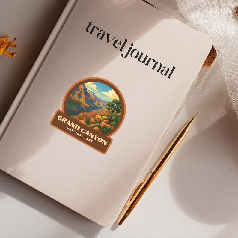 May include: A cream-colored travel journal with the words "travel journal" printed in black. A Grand Canyon National Park sticker is affixed to the cover. A gold pen rests on the journal. The sticker features a landscape of the Grand Canyon.