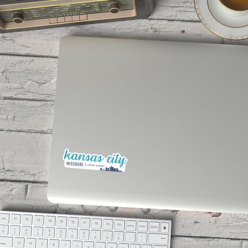 Puede incluir: Una pegatina con un dise&ntilde;o azul y blanco que dice "kansas city MISSOURI | 39.0997&deg; N, 94.5786&deg; W" con una silueta de horizonte.