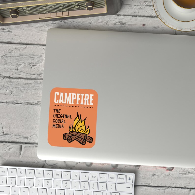 May include: An orange sticker with the word "CAMPFIRE" in white block letters. Below the word "CAMPFIRE" is the text "THE ORIGINAL SOCIAL MEDIA". A graphic of a campfire with flames and logs is below the text.