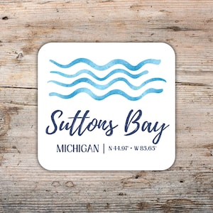 May include: White square coaster with rounded corners. The coaster features blue watercolor waves above the text "Suttons Bay, Michigan" and coordinates. The design is on a wooden background.
